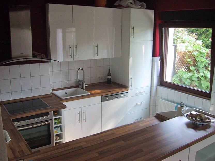 Küche nach Maß Moderne Einbauküche in Hochglanzoptik mit weißen Fronten, Holzarbeitsplatte, Einbauherd und großem Fenster mit Gartenblick.