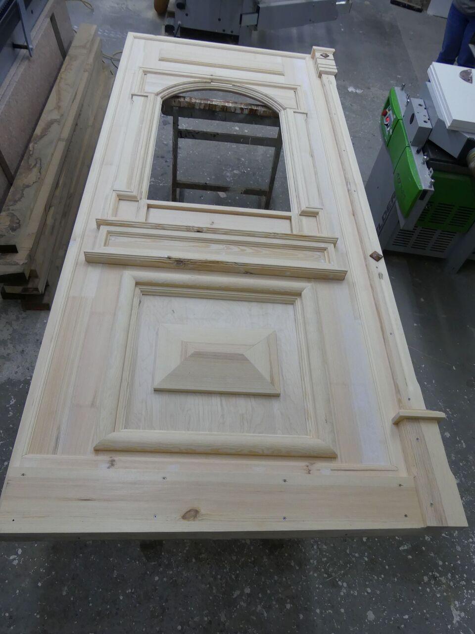 Unbehandelte massive Holztür mit aufwendigen Profilen und Ausschnitt für Glas während der Fertigung in der Werkstatt.